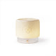 A ceramic electric wax warmer with a ribbed, flared top section in a warm cream color that glows with a soft yellow light, resting on a white cylindrical base with a small gold button.