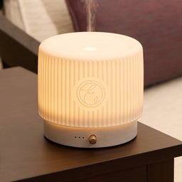 A cylindrical LED diffuser and humidifier with a ribbed white body, glowing warm yellow light, and a small gold button on the base, placed on a dark wooden table in a cozy indoor setting.