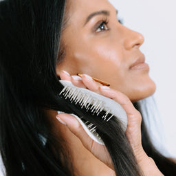 A close-up lifestyle shot of a woman with long, straight black hair using a white and gold hairbrush to brush her hair against a plain white background.