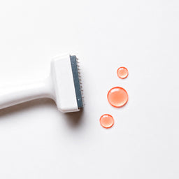 A close-up of a white pet grooming brush with a dark gray metal edge, positioned next to three clear droplets of orange liquid on a clean white background.
