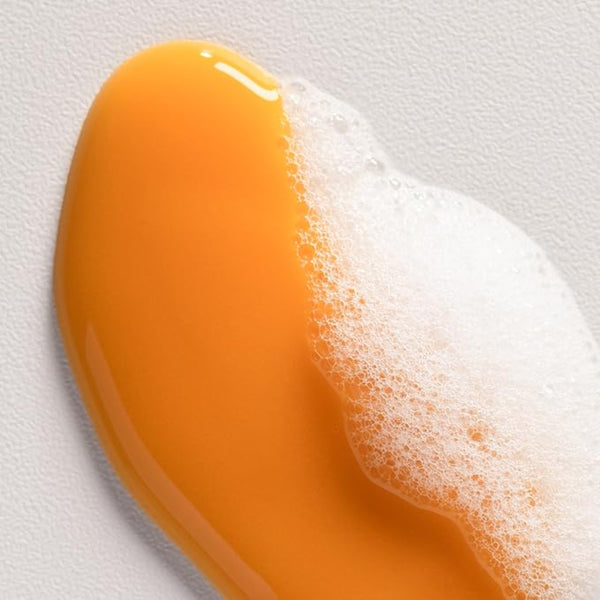 A close-up of a glossy, translucent orange liquid with white foam on a textured light gray background.