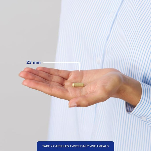 A person in a light blue and white striped shirt holds an open hand showing a single capsule pill, with a white line and blue text indicating its size is 23 mm. A dark blue banner at the bottom reads 'TAKE 2 CAPSULES TWICE DAILY WITH MEALS'.