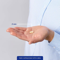 A hand holding a small beige capsule with a measurement label reading 2 mm, shown against a person's torso in a light blue striped shirt. Text at the bottom says TAKE 1 CAPSULE DAILY WITH A MEAL.