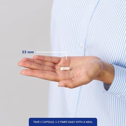 A person's hand holding a white capsule pill, with a measurement overlay indicating its size is 23 mm. The person is wearing a light blue and white striped shirt against a light gray background, with a blue text banner at the bottom providing dosage instructions.