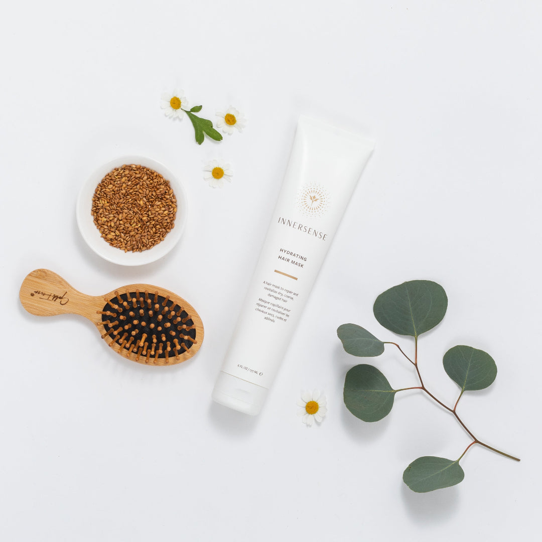 A white tube of INAYUSHI shampoo is displayed in a styled flat-lay arrangement on a white background, surrounded by a wooden hairbrush, a small bowl of brown seeds, and eucalyptus leaves.