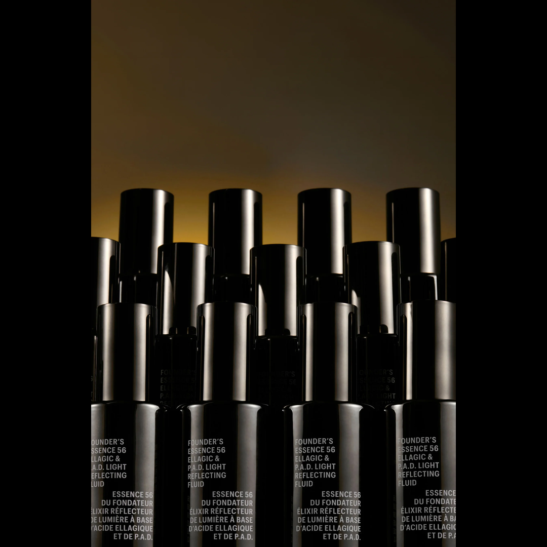 A group of black cosmetic bottles with silver caps arranged in a row against a dark background with a warm golden glow near the top. The bottles have white text on the lower section that reads 'BUNDER'S ESSENCE 56 LUMIÈRE & LUMIÈRE REFLECTING'.