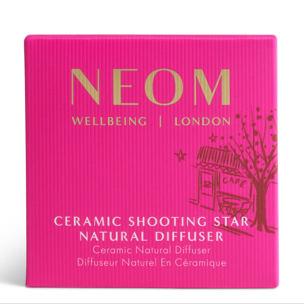 Bright pink square product box for a NEOM Wellbeing London Ceramic Shooting Star Natural Diffuser, featuring gold and white text and a small cafe-and-tree illustration on a white background.