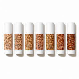 Six white cosmetic tubes arranged in a row on a white background, each showing a different shade of tan and brown seed beads in the clear center window.
