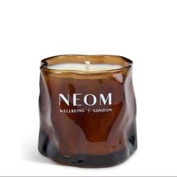 A NEOM Wellbeing London scented candle in a wavy, organic-shaped amber glass jar, shown on a clean white background.