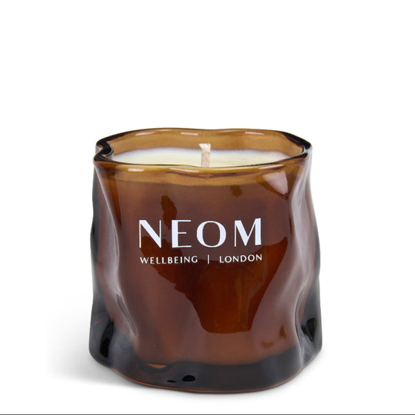 A NEOM Wellbeing London scented candle in a wavy, organic-shaped amber glass jar, shown on a clean white background.