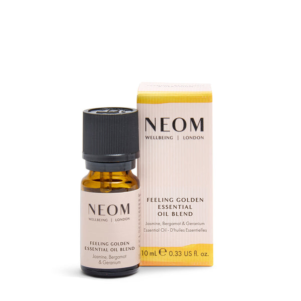 NEOM Wellbeing London 'Feeling Golden' essential oil blend shown as a small amber glass bottle with a black cap next to its cream and yellow box, on a white background with a beige banner at the bottom reading 'PRECIOUS OILS'.