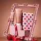 Gatineau Golden Glow body care set displayed in front of an ornate pink picture frame, featuring a body wash tube, a moisturizer tube, and a patterned box with a red ribbon and red ornament in the foreground.