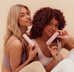 Two women holding pink lip products against a beige background