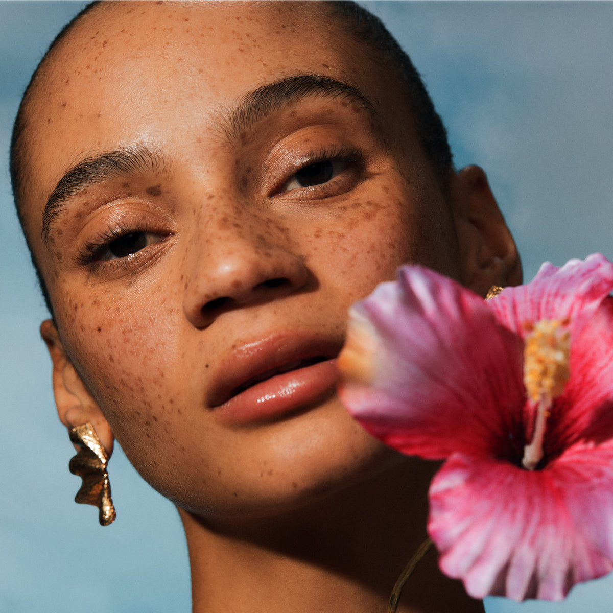 Close-up of a person with a pink flower in front of their face against a blue sky.