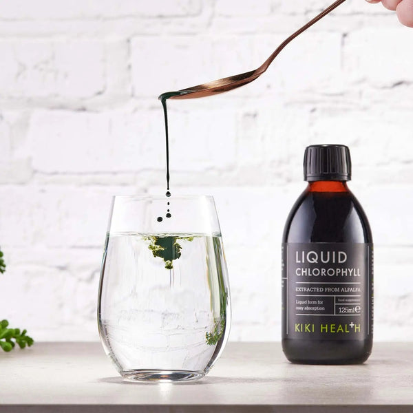 Bottle of KIKI Health Liquid Chlorophyll being poured into a glass with a white brick wall background.