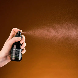Hand holding a bottle of magnesium oil spray against a brown background