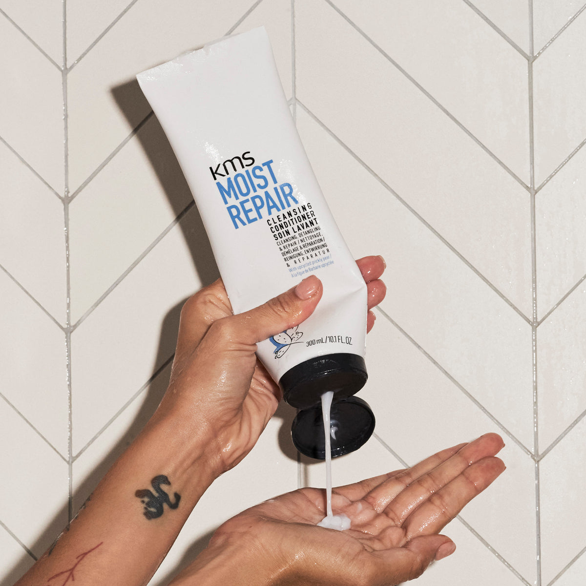 Hand holding a tube of KMS Moist Repair product with a tiled bathroom background
