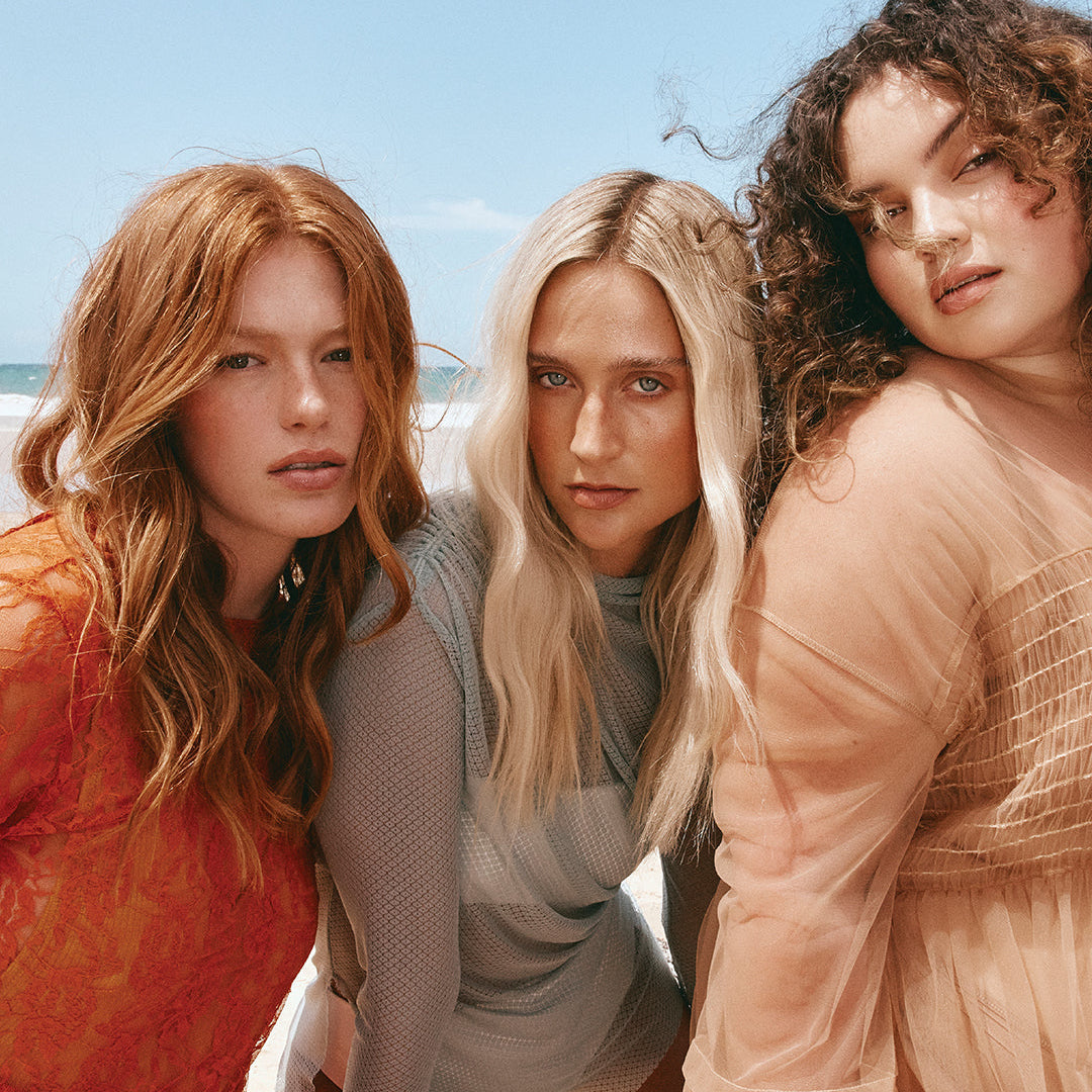 Three women standing together on a beach with clear blue sky.
