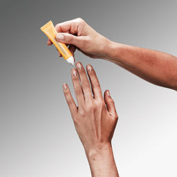 Hand applying a yellow tube of cream to another hand against a gray background