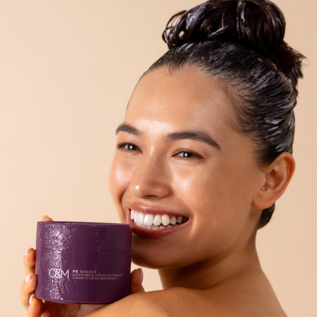 A smiling woman with dark hair in a bun holds a purple O&M Paris Masque jar next to her face against a warm beige background.