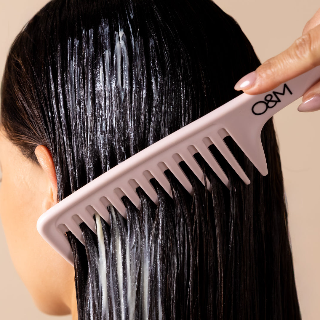 A close-up of a person with wet, dark hair being combed with a wide-tooth pink comb. A hand with manicured nails is visible holding the comb, set against a plain beige background.