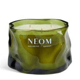 A three-wick scented candle in a wavy, organic-shaped translucent olive green glass container, with 'NEOM WELLBEING | LONDON' printed in white on the front, set against a plain white background.