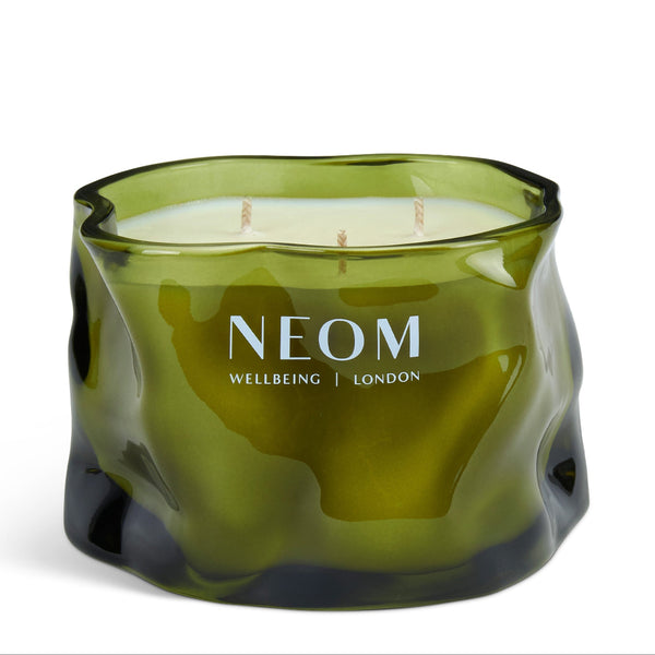 A three-wick scented candle in a wavy, organic-shaped translucent olive green glass container, with 'NEOM WELLBEING | LONDON' printed in white on the front, set against a plain white background.