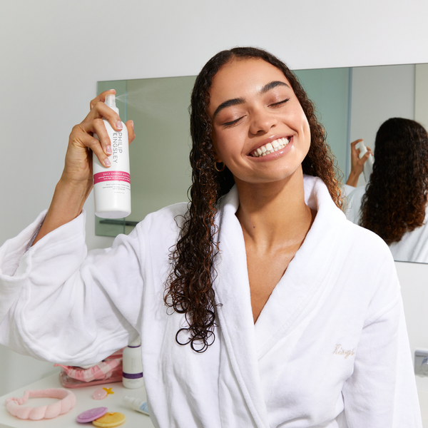 A woman in a white bathrobe smiles while holding up a white bottle of hair product with pink accents in front of a bathroom mirror.