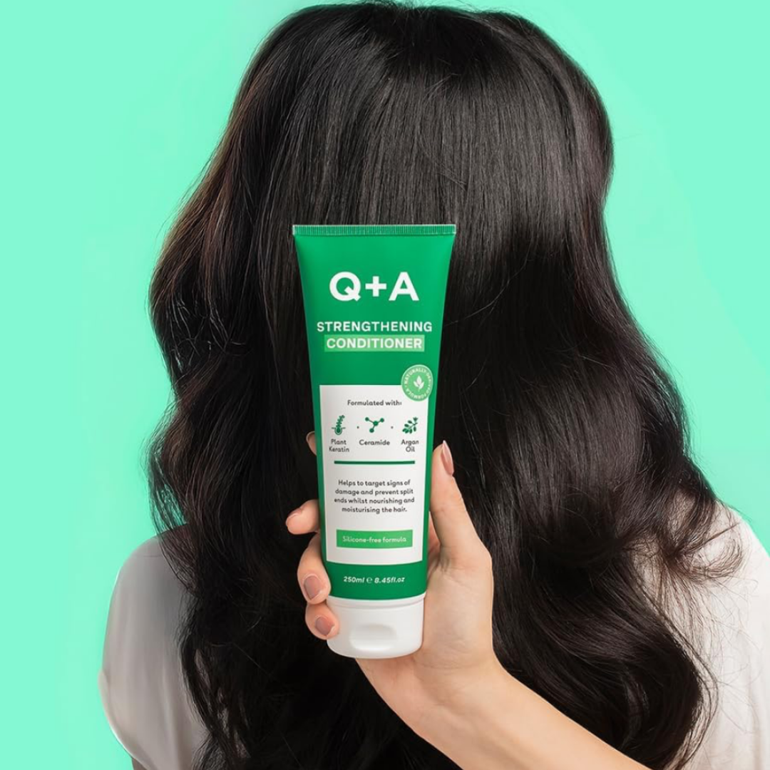 Person holding a Q+A Strengthening Conditioner tube with long dark hair against a green background