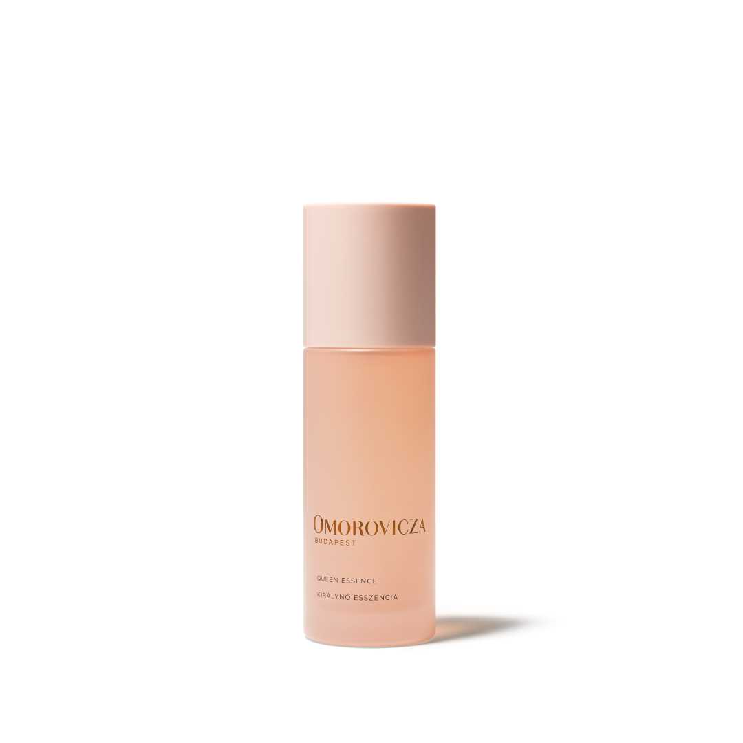 A bottle of Omorovicza Queen Essence Facial Essence in a soft peach-pink shade, featuring a matching pale pink cap, set against a clean white background with a soft shadow.