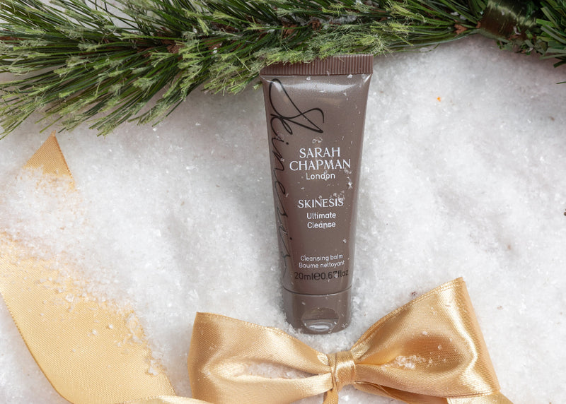 Tube of Sarah Chapman skincare product on a snowy surface with a gold bow and pine branches.