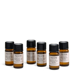 Six amber glass essential oil bottles with black ribbed caps and cream labels, arranged in two rows on a white background. The labels show the brand NEOM and various scent names including 'Cozy Nighter Essential Oil Blend' and 'Sparkling Joy Essential Oil Blend'.