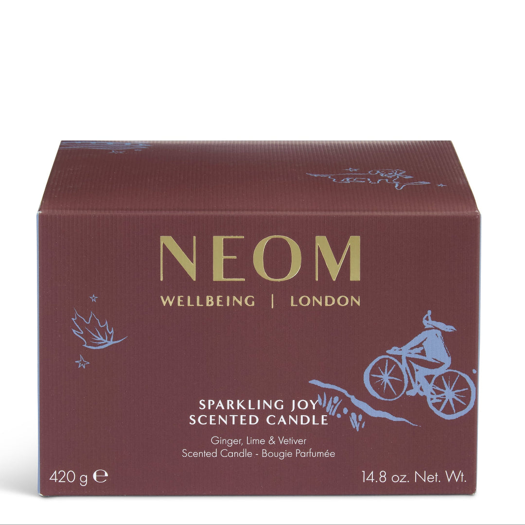 A rectangular NEOM Wellbeing London scented candle box in a deep burgundy-brown color, featuring gold lettering and light blue line-art illustrations of a cyclist and botanical motifs, set against a plain white background.