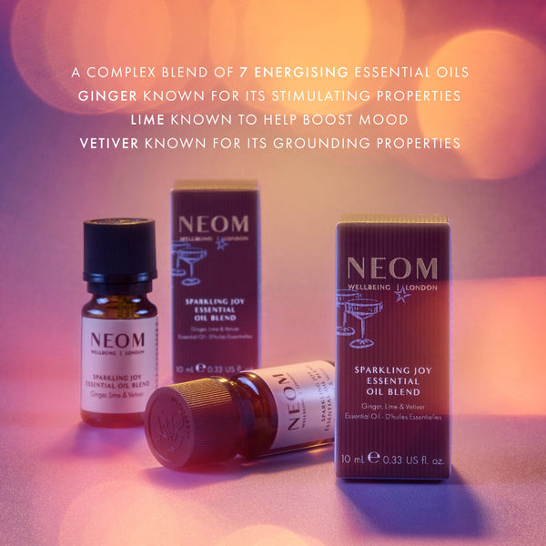 NEOM Sparkling Joy Essential Oil Blend set with amber glass bottles and matching dark brown boxes, arranged on a softly lit pink-purple surface with decorative bokeh. White text at the top describes the blend's ingredients: ginger, lime, and vetiver.