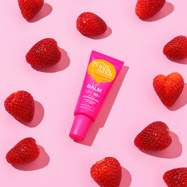 A pink squeeze tube of Bondi Sands Strawberry Lip Balm SPF 50+ lies diagonally on a pink background, surrounded by fresh red strawberries.