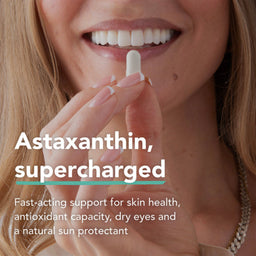 Close-up of a smiling woman holding a white capsule pill between her fingers, with text reading 'Astaxanthin, supercharged' and additional product benefits on a light background.