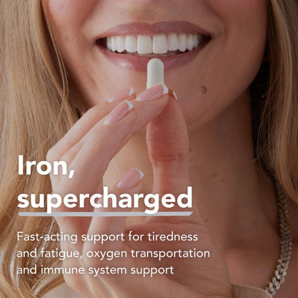 Close-up of a smiling woman holding a white tablet near her mouth, with text reading 'Iron, supercharged' and additional supplement benefit copy on a light background.