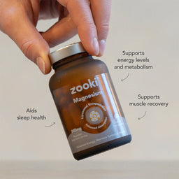 A hand holds a brown amber bottle of Zooki Magnesium supplement. The bottle has a silver cap and a label with white and gold text.