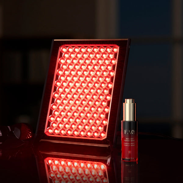 A red LED light therapy panel glowing brightly next to a red and gold skincare bottle on a reflective dark surface.