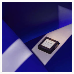 A small rectangular product box with a white label sits on a geometric blue and white surface. The box appears dark, likely black or very deep navy, with a textured pattern and a visible 'L'Or élixir' label.