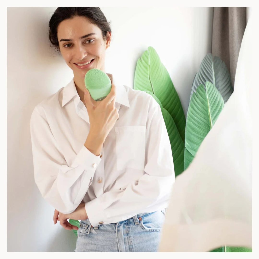 A woman wearing a white button-up shirt and light blue jeans holds a light green oval cosmetic container near her face, standing against a white wall with large green leaves in the background.