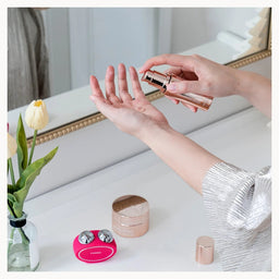 A person's hands apply a cosmetic product from a rose gold tube onto their palm, with other cosmetic items and a vase of tulips on a white vanity table in front of a mirror.