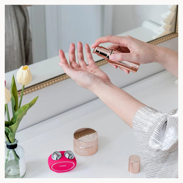 A person's hands apply a cosmetic product from a rose gold tube onto their palm, with other cosmetic items and a vase of tulips on a white vanity table in front of a mirror.