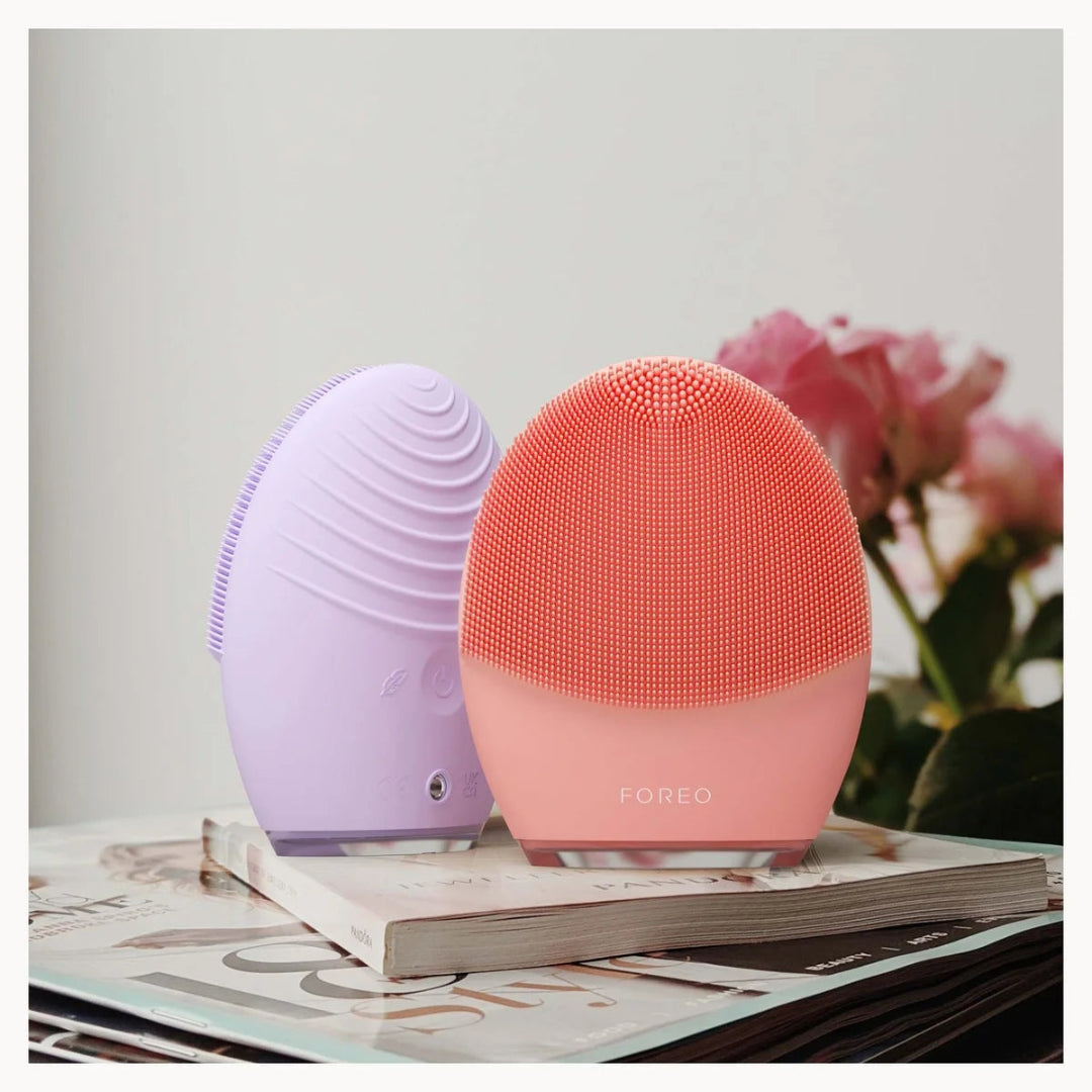 Two FOREO facial cleansing devices, one in lavender purple and one in coral pink, displayed on a book with blurred pink flowers in the background.
