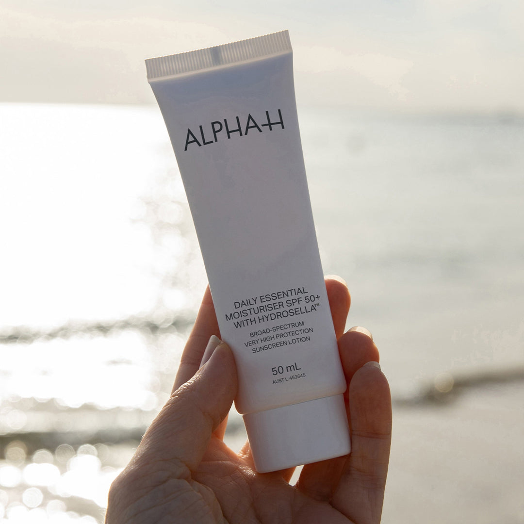 Hand holding a tube of Alpha-H daily essential moisturizer with a blurred beach background