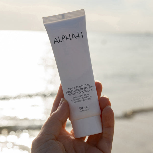 Hand holding a tube of Alpha-H daily essential moisturizer with a blurred beach background