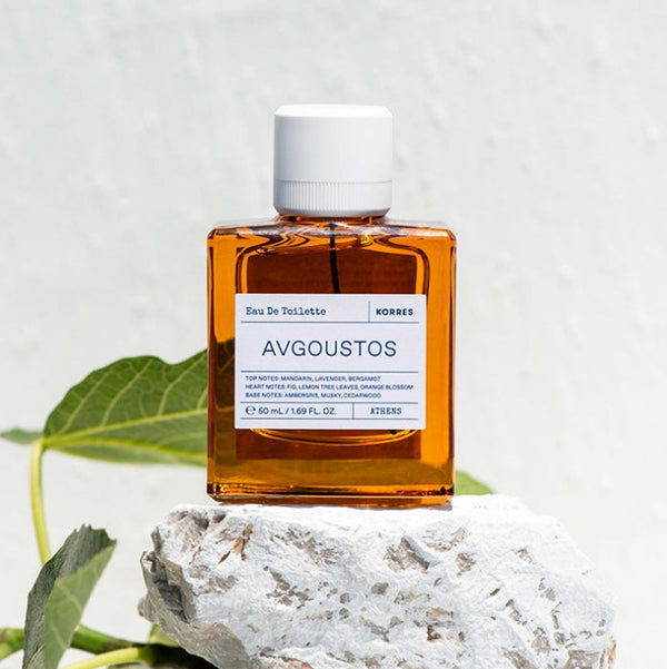 A rectangular amber glass bottle of Augoustos Eau De Toilette with a white cap and white label, styled on a white stone with green leaves against a textured white background.